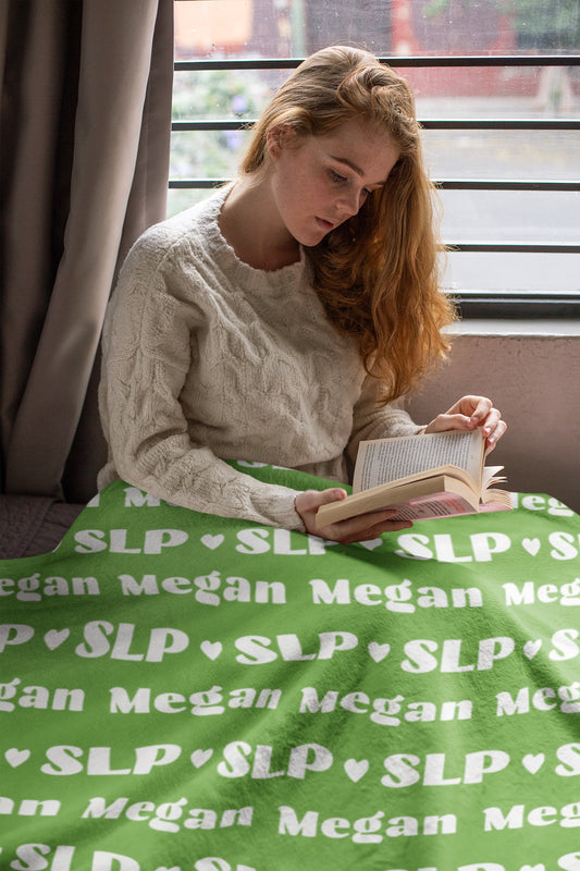 Woman reading book by window with green plush blanket that says SLP and her name Megan across the whole blanket in retro font